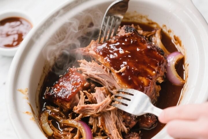 Overhead shot of slow-cooked BBQ pulled pork being shredded in the crockpot with two forks, glisteni